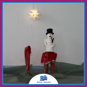 Auf zwei kurzen roten Rohren am unteren Bildrand steht eine Schneemannfigur vor einer hellen Wand. Ein weiteres Rohr ragt daneben aus dem Boden. Der Boden ist mit Folie belegt. Ein kleiner Stern hängt über dem Schneemann.